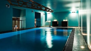 Una amplia piscina interior con una decoración azul, una escalera de madera y suelo de baldosas.