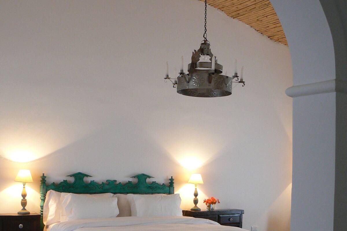 A bedroom with a bed, two bedside tables with lamps, a bench, and a hanging chandelier.