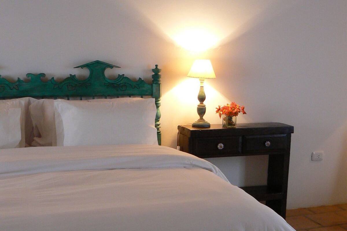 A neatly made bed with white linens, a green headboard, a bedside table with a lamp and flowers, and a small nightstand.