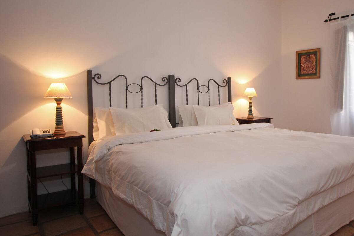 A neatly made bed with white linens, two bedside tables with lamps, a framed picture on the wall, and a window with sheer curtains.