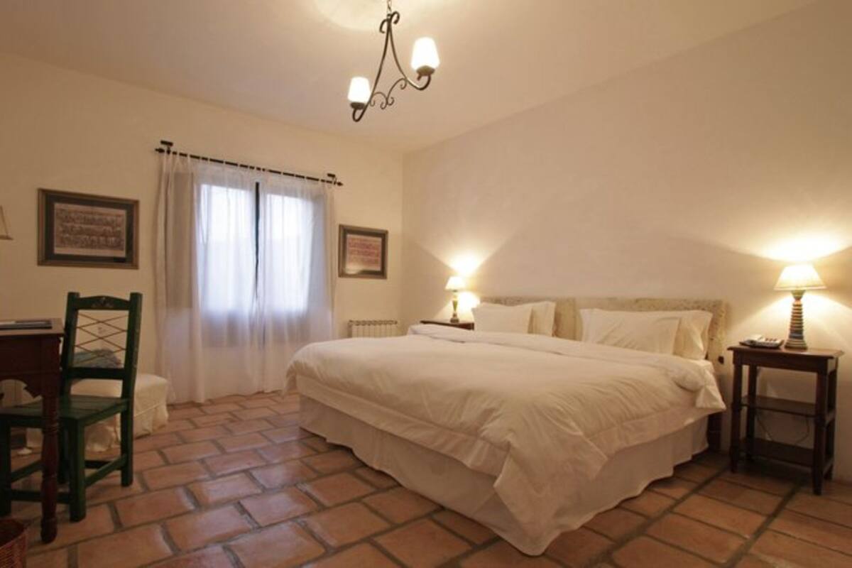 A bedroom with a large bed, two bedside tables with lamps, a chair, a desk, a radiator, and a window with curtains.
