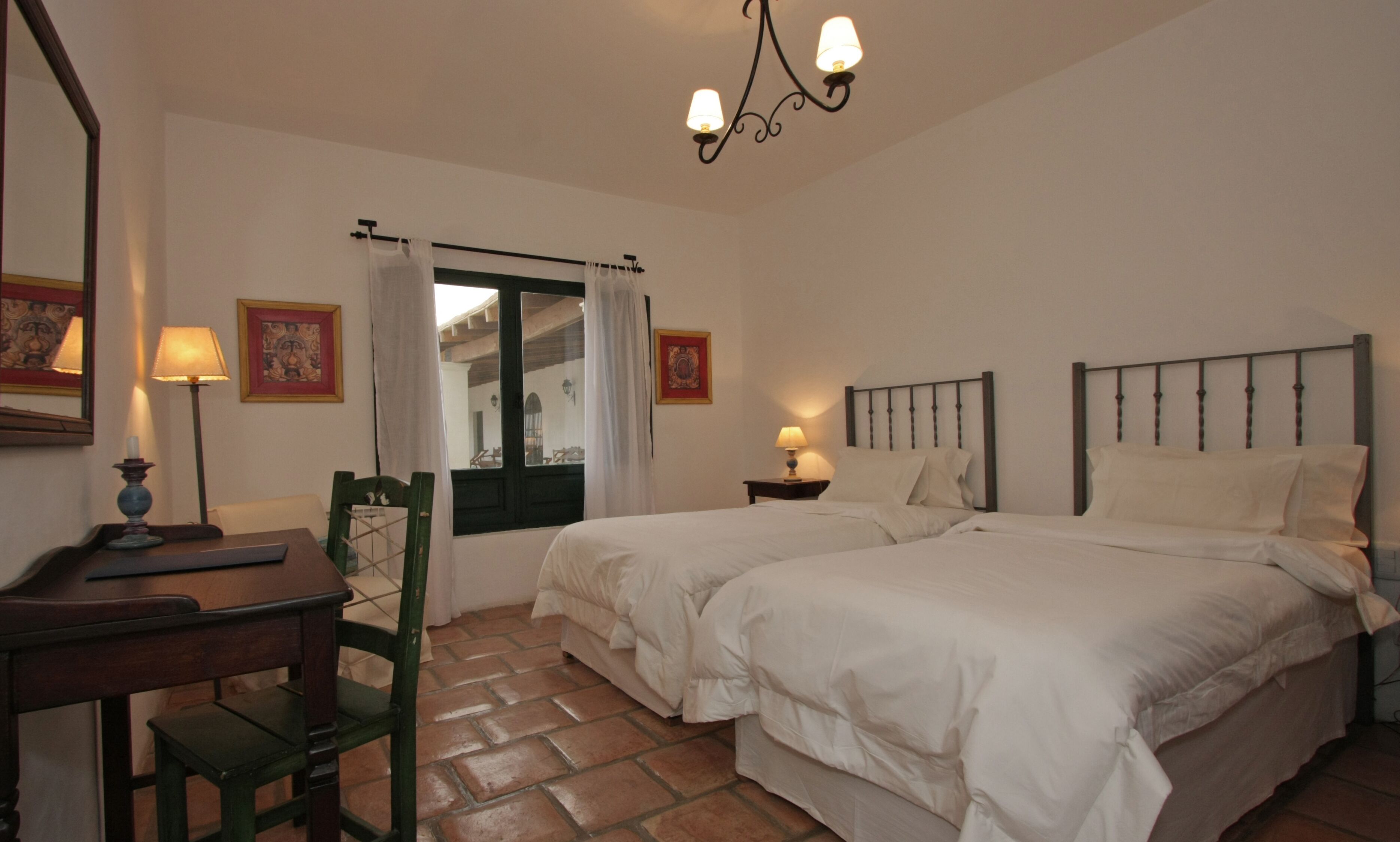 A bedroom with two beds, a desk, a chair, a window with curtains, and a chandelier.
