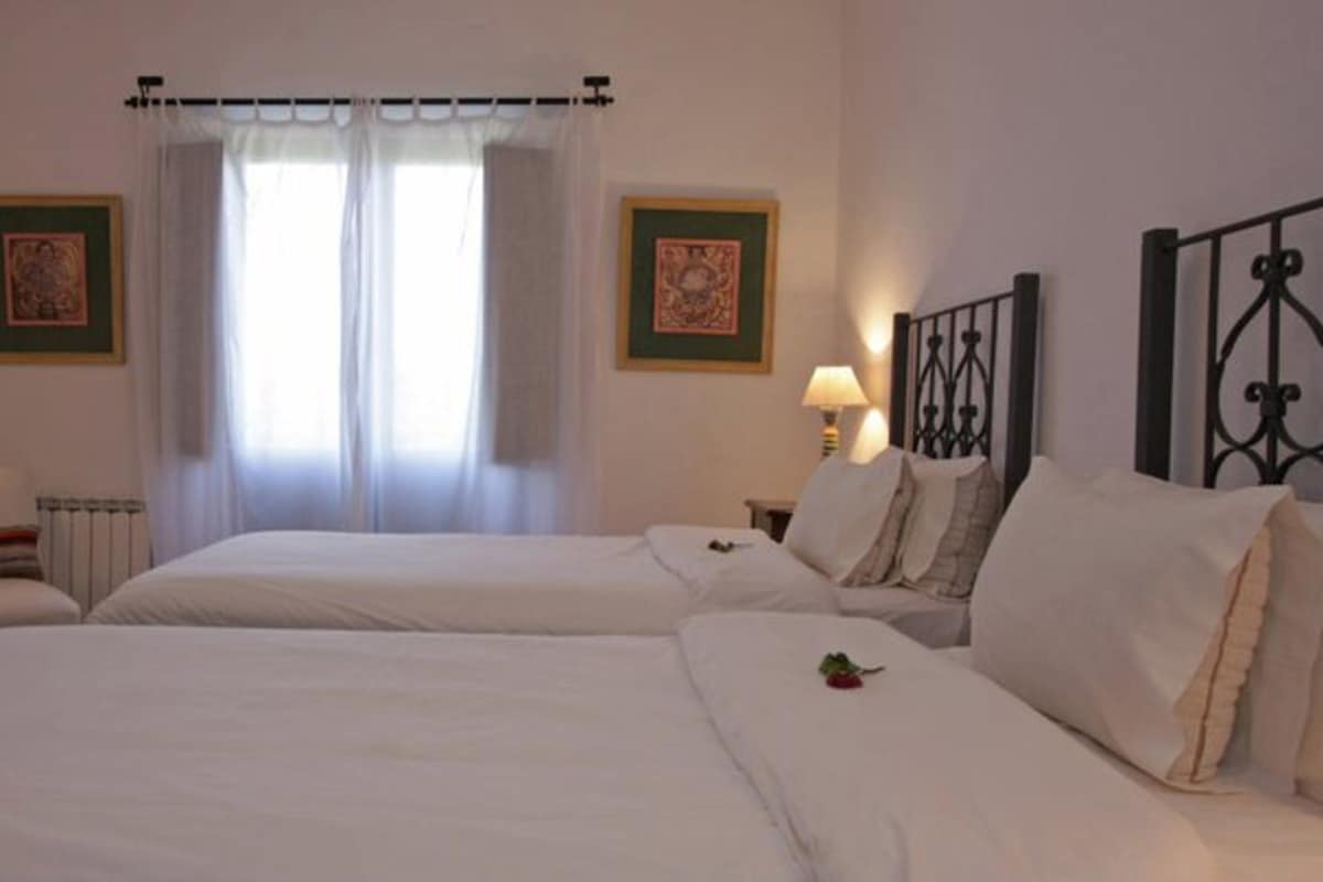 Habitación doble con dos camas, un radiador, una silla, dos mesitas de noche con lámparas y dos cuadros en las paredes.