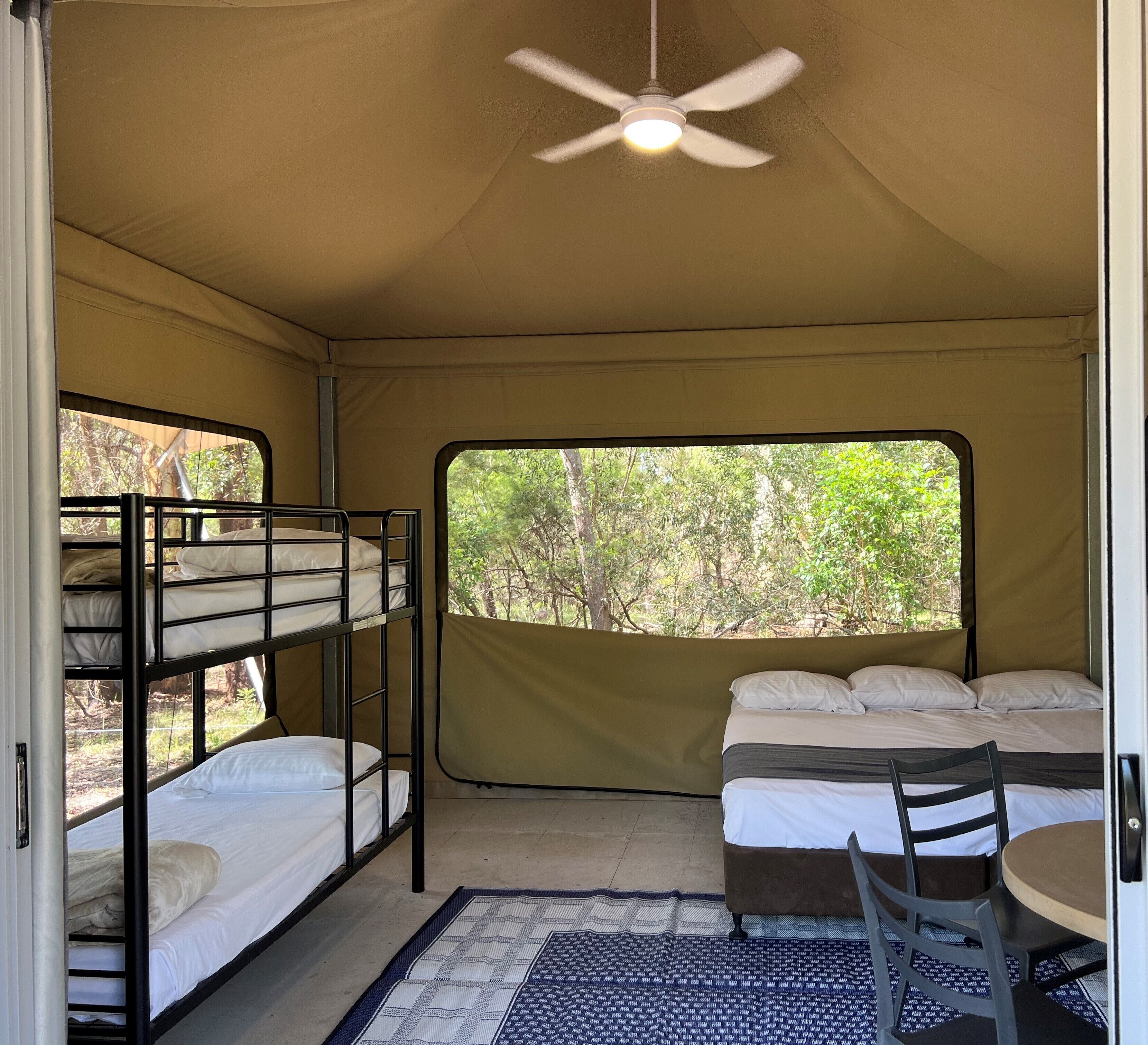 A tent with bunk beds, a ceiling fan, a table with chairs, and a view of trees through a window.