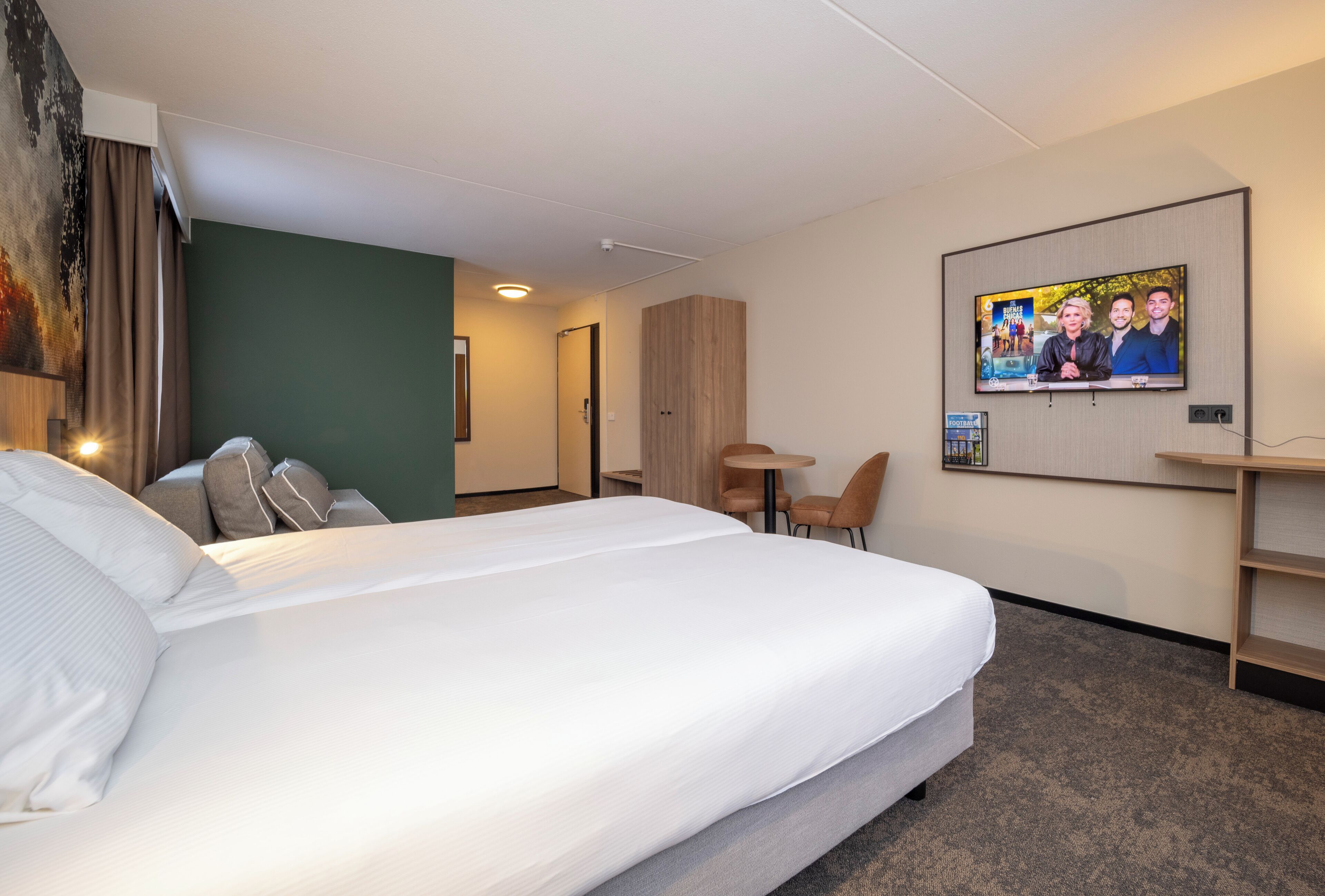 A hotel room with a large bed, a desk, a chair, a TV mounted on the wall, and a painting on the wall.