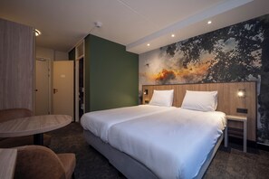 A hotel room with a large bed, a round table, and a chair. There is a painting on the wall and a door leading to another room.