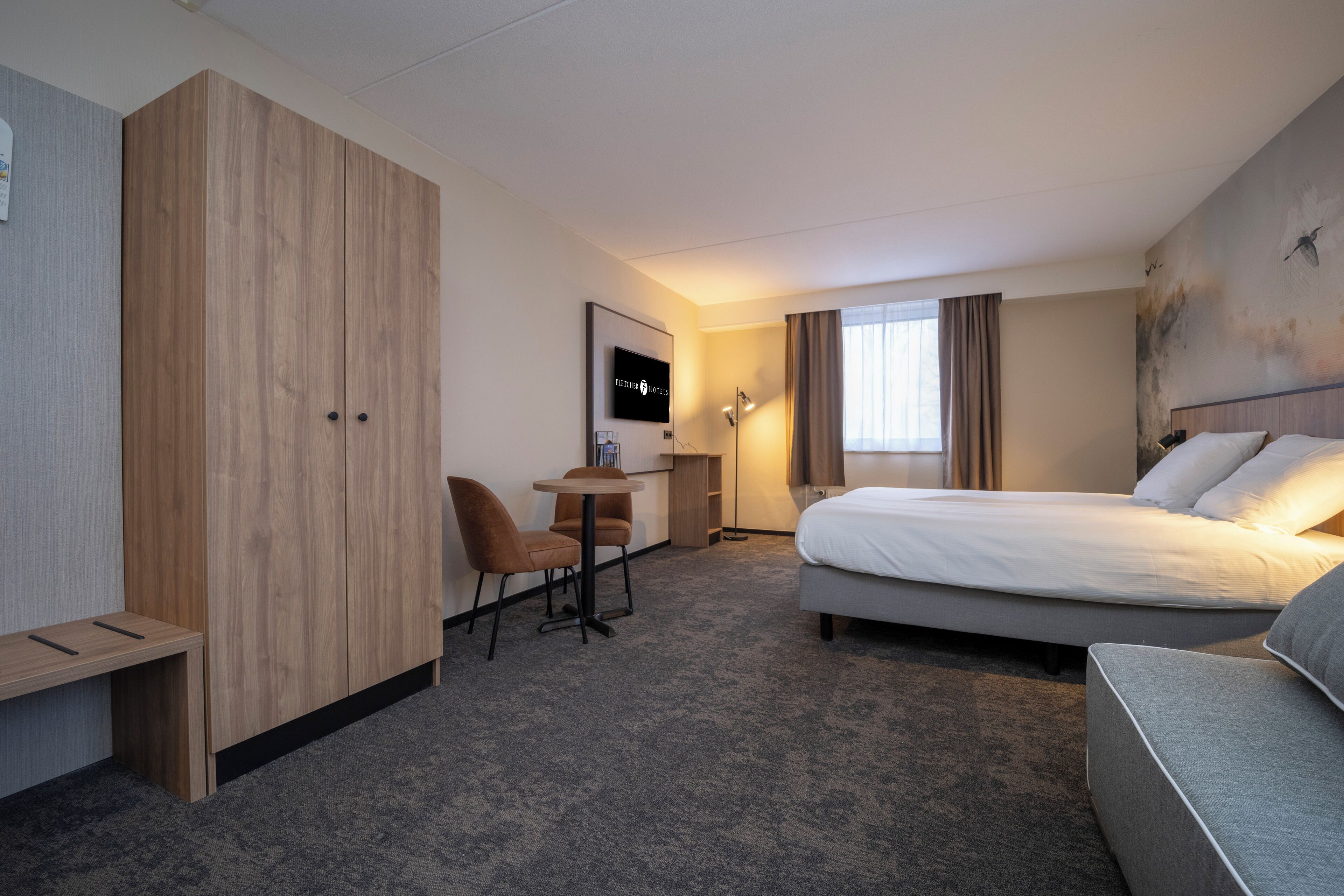 Habitación de hotel con cama, escritorio, silla, armario y un amplio ventanal con cortinas.