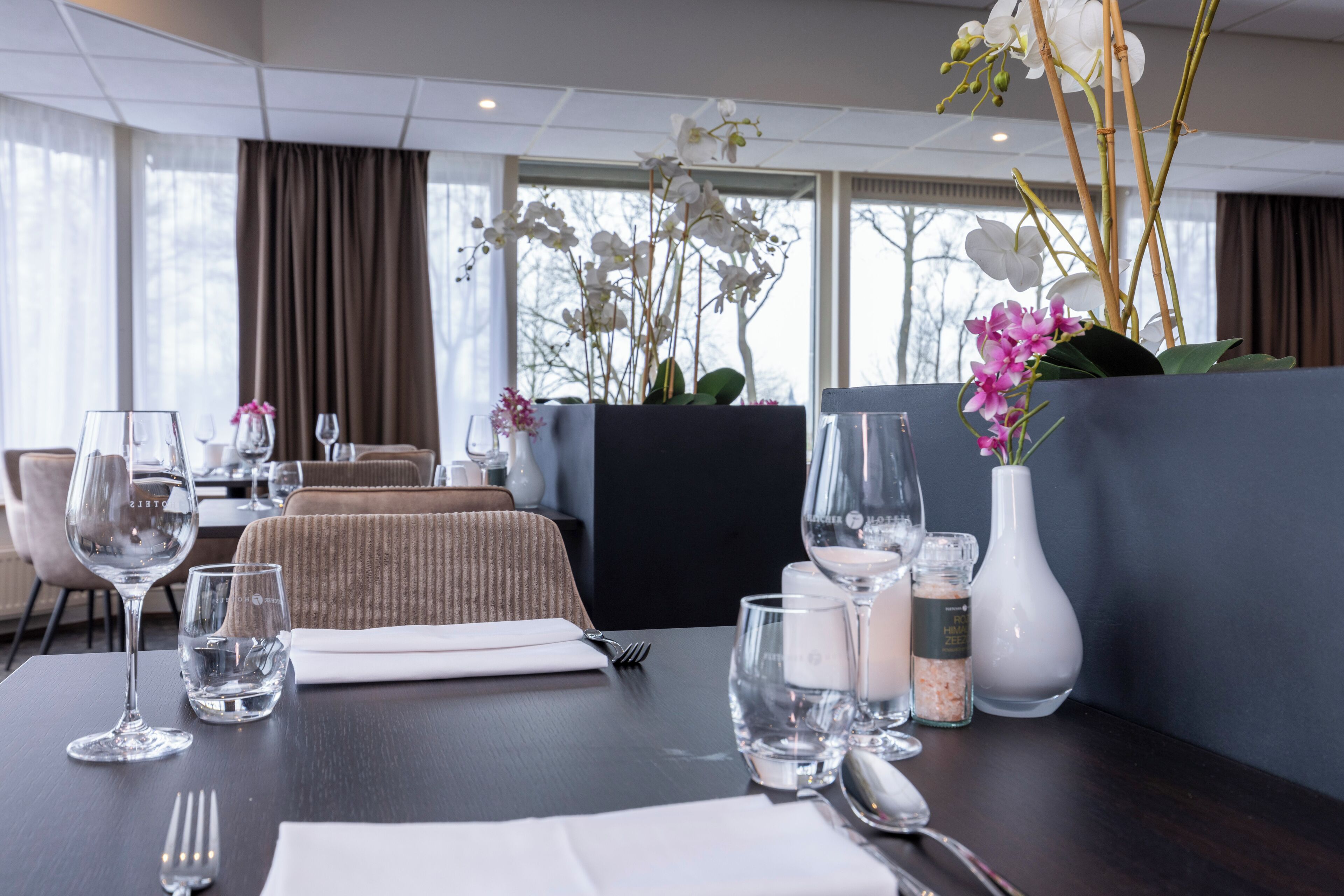 Een eettafel, gedekt met helder glaswerk, een witte vaas en een klein flesje, met uitzicht op een restaurant en een raam met gordijnen.