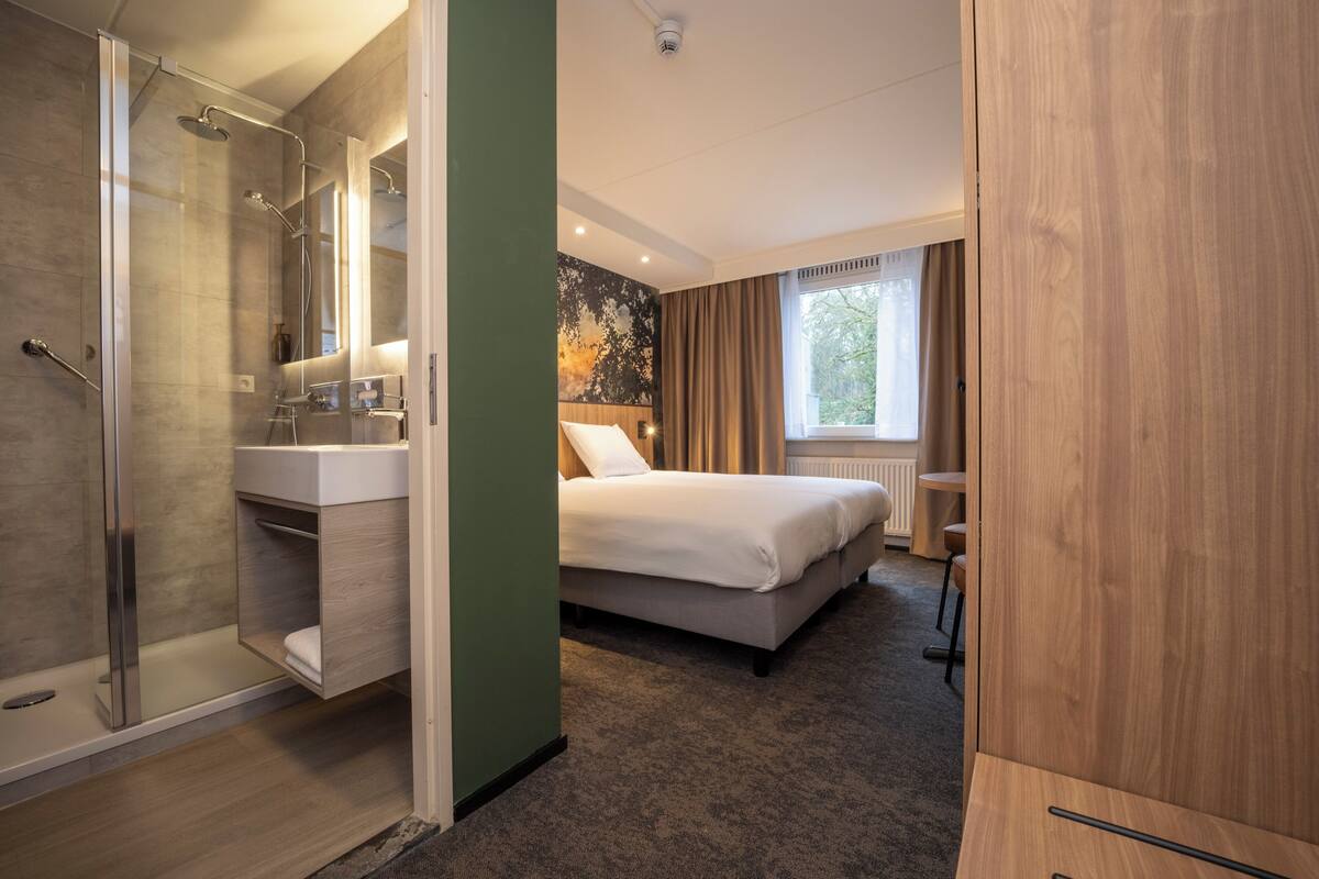A modern hotel room with a glass-enclosed shower, a large bed with white linens, a wooden wardrobe, and a view of trees through a window.