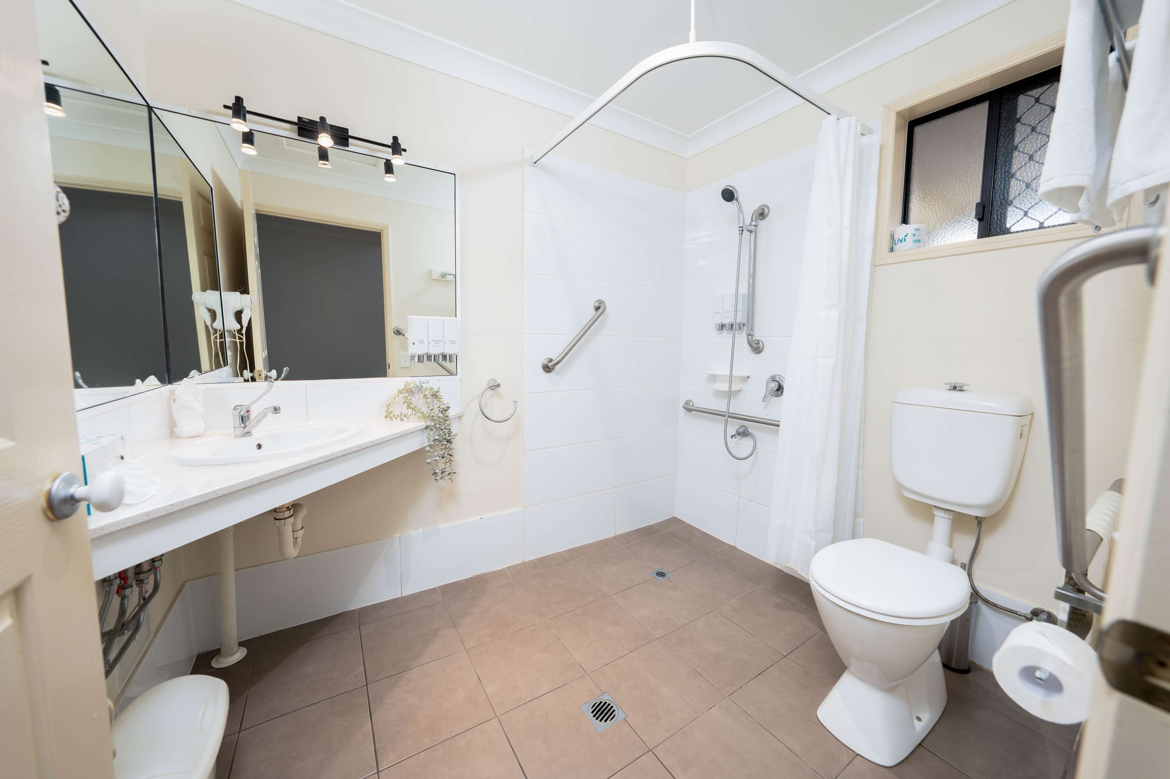 Un baño moderno con lavamanos, espejo y retrete blancos. Incluye zona de ducha con barras de apoyo y ventana con una toalla colgada.