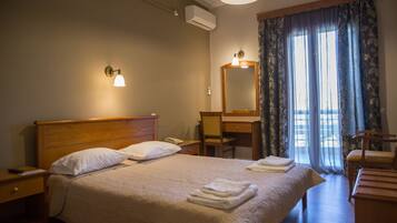 A hotel room with a bed, bedside table, lamp, mirror, and a view of the outdoors through a window with curtains.