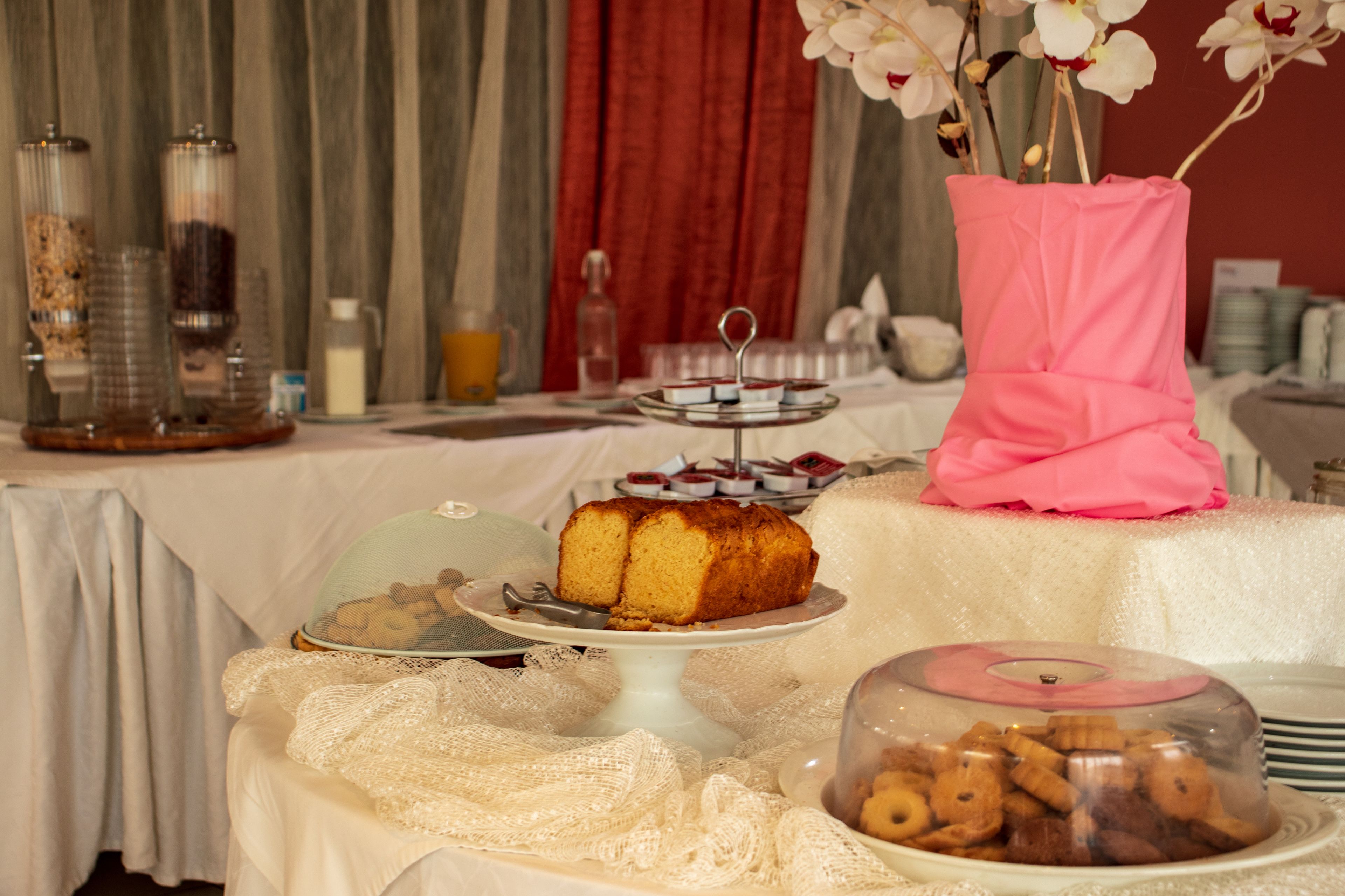 A buffet table with various food items, including a cake and pastries, set against a backdrop of red curtains and a floral arrangement.