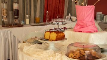 A buffet table with various food items, including a cake and pastries, set against a backdrop of red curtains and a floral arrangement.
