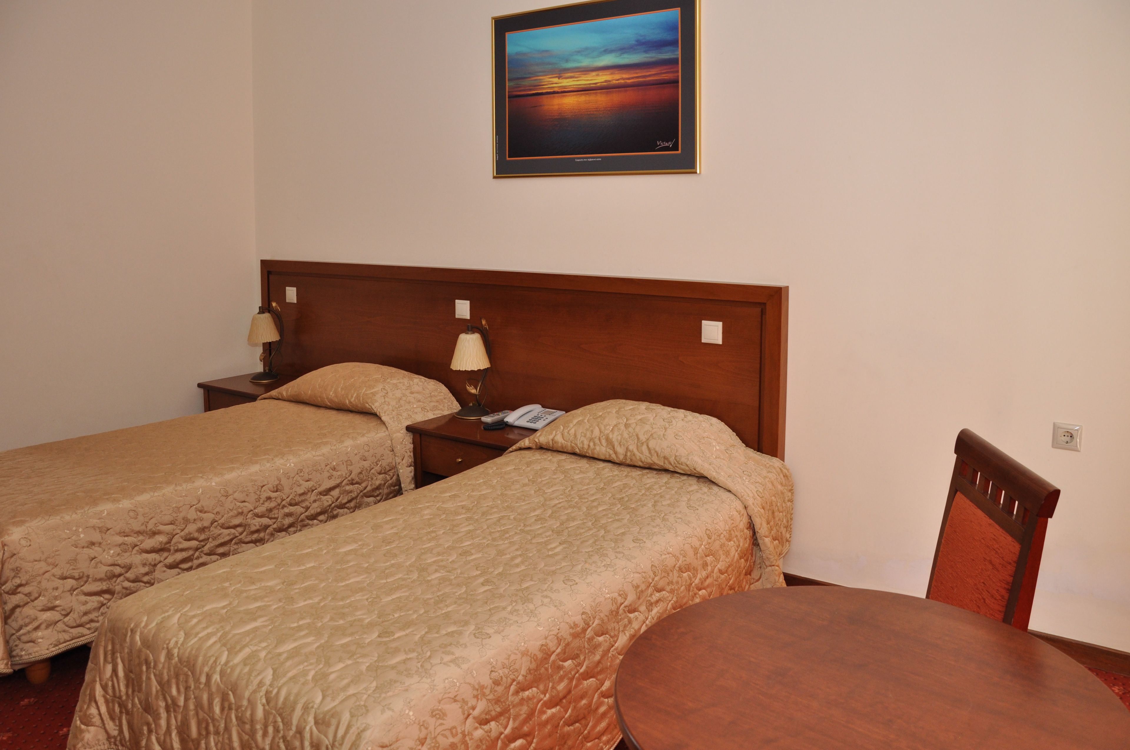 A hotel room with two single beds, a wooden headboard, a bedside table with a lamp, a phone, and a painting of a sunset on the wall.
