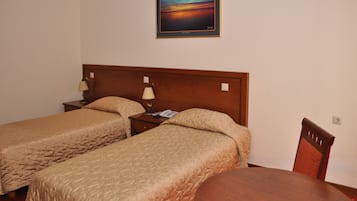 A hotel room with two single beds, a wooden headboard, a bedside table with a lamp, a phone, and a painting of a sunset on the wall.