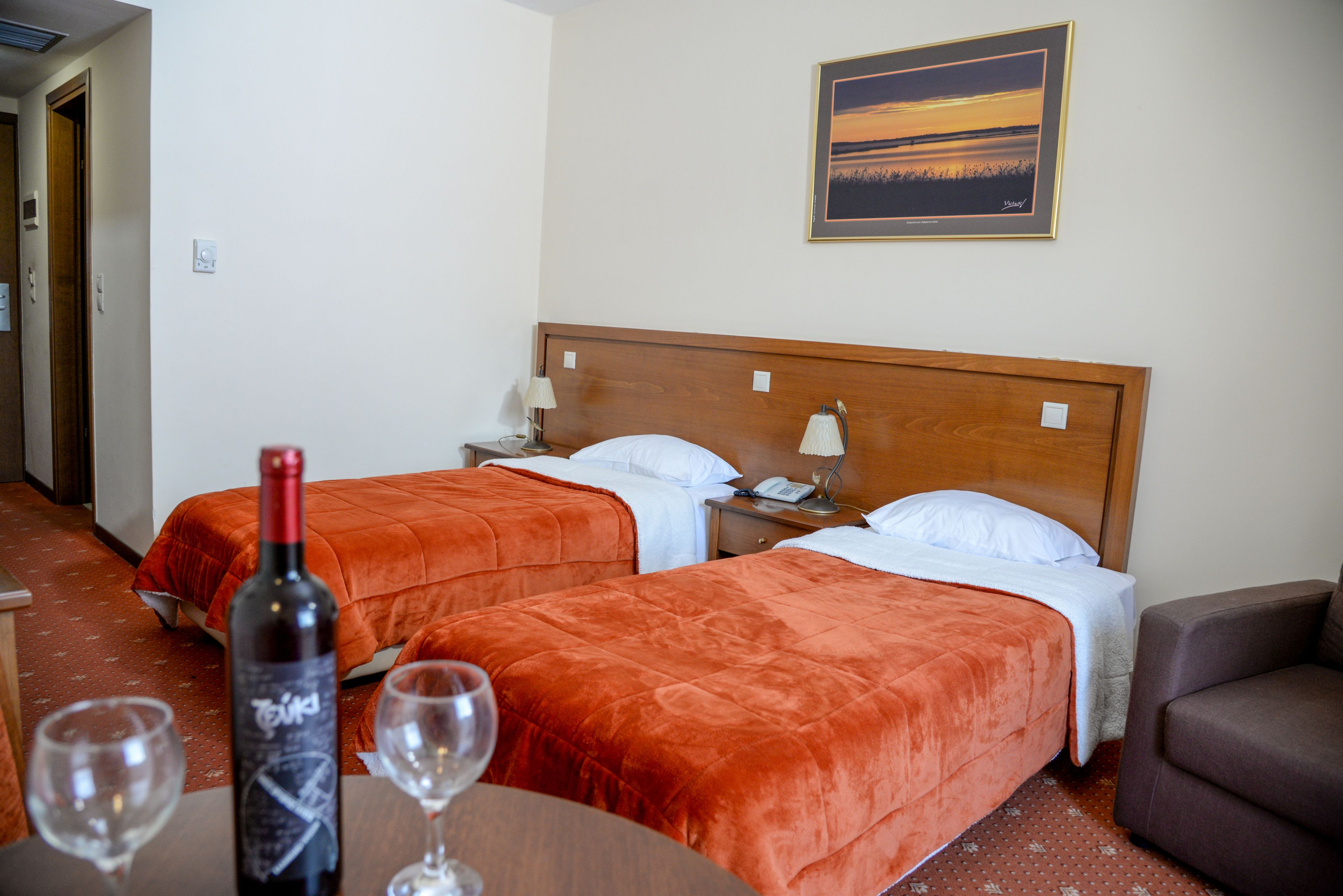 A hotel room with two beds, a wooden headboard, a painting on the wall, and a bottle of wine on a table.