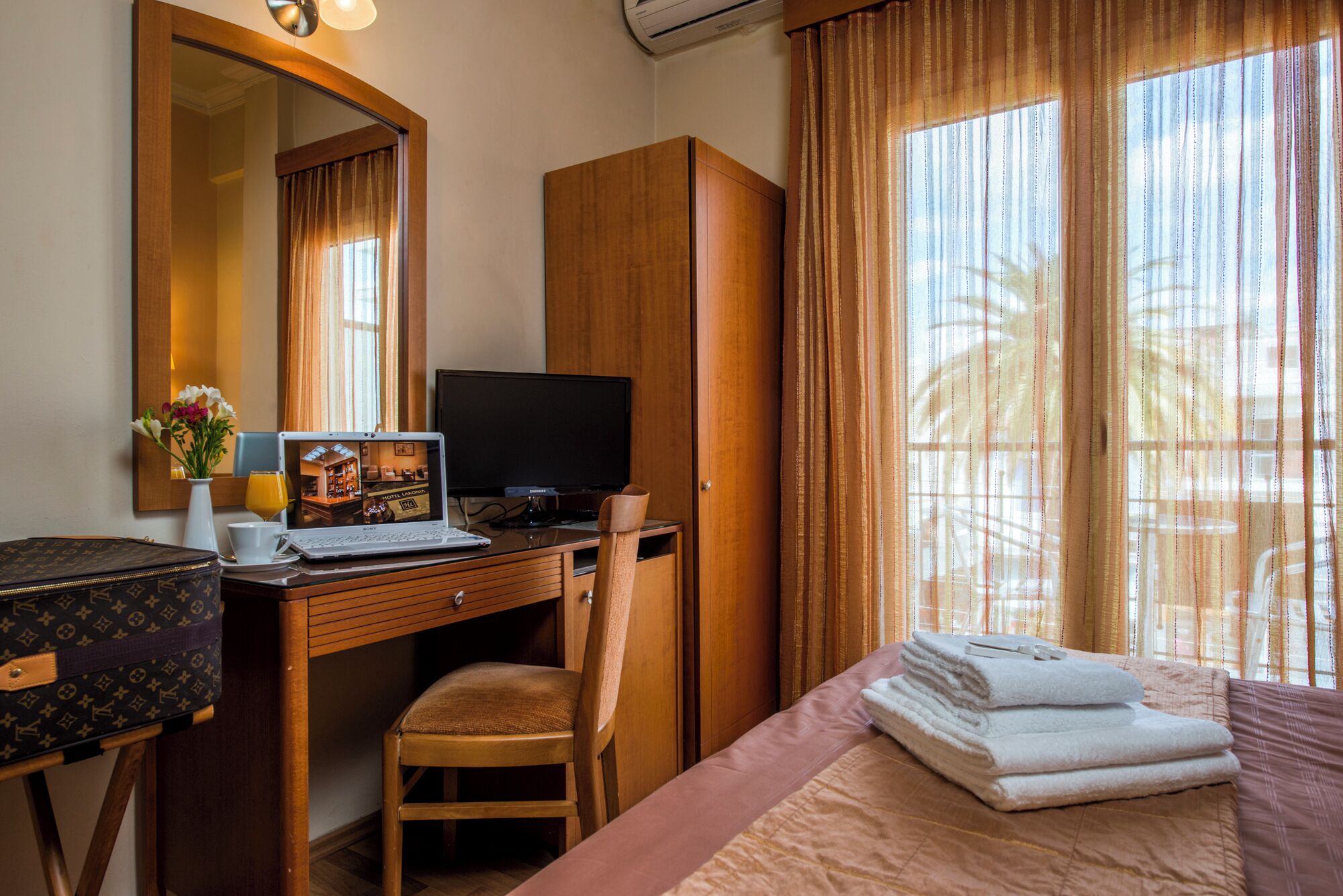 Habitación de hotel con escritorio de madera, laptop, televisor, silla, cama con cubrecama rosa y beige, y ventana con cortinas.