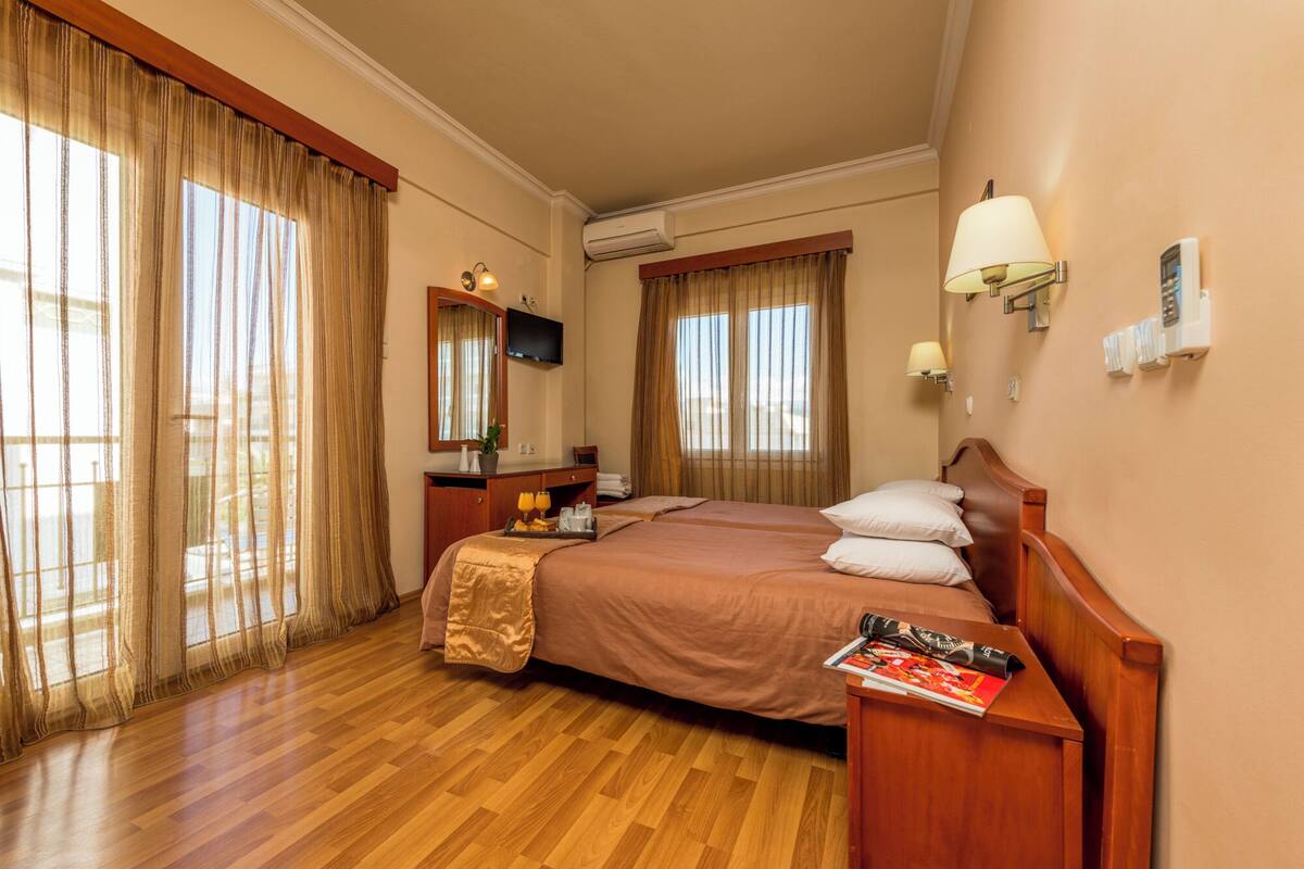 Habitación de hotel con cama de madera, mesita de noche con una revista, espejo, televisor y ventana con cortinas.