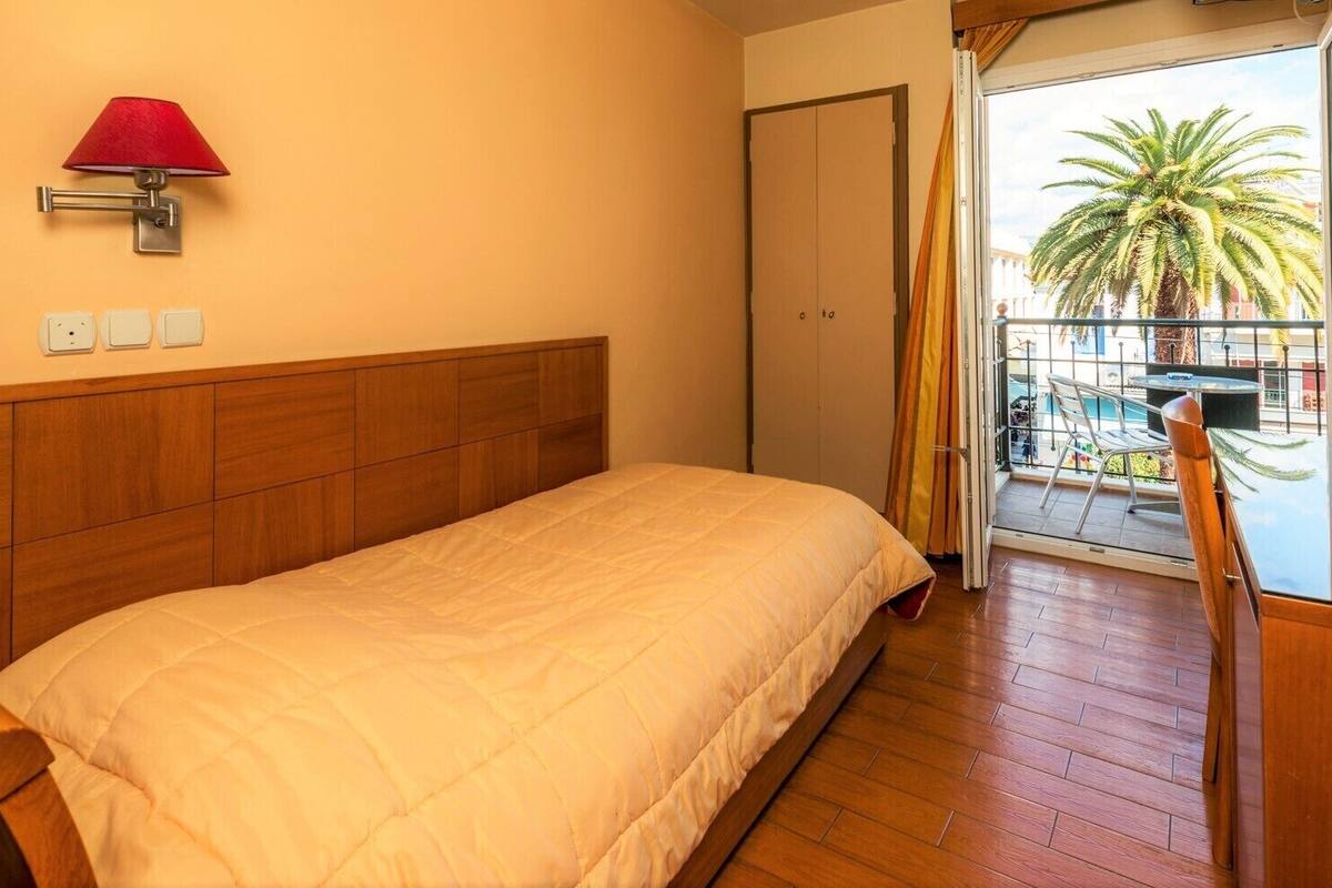 Habitación de hotel con cabecera de madera, cama con edredón blanco, lámpara roja y vista a un balcón con una palmera.