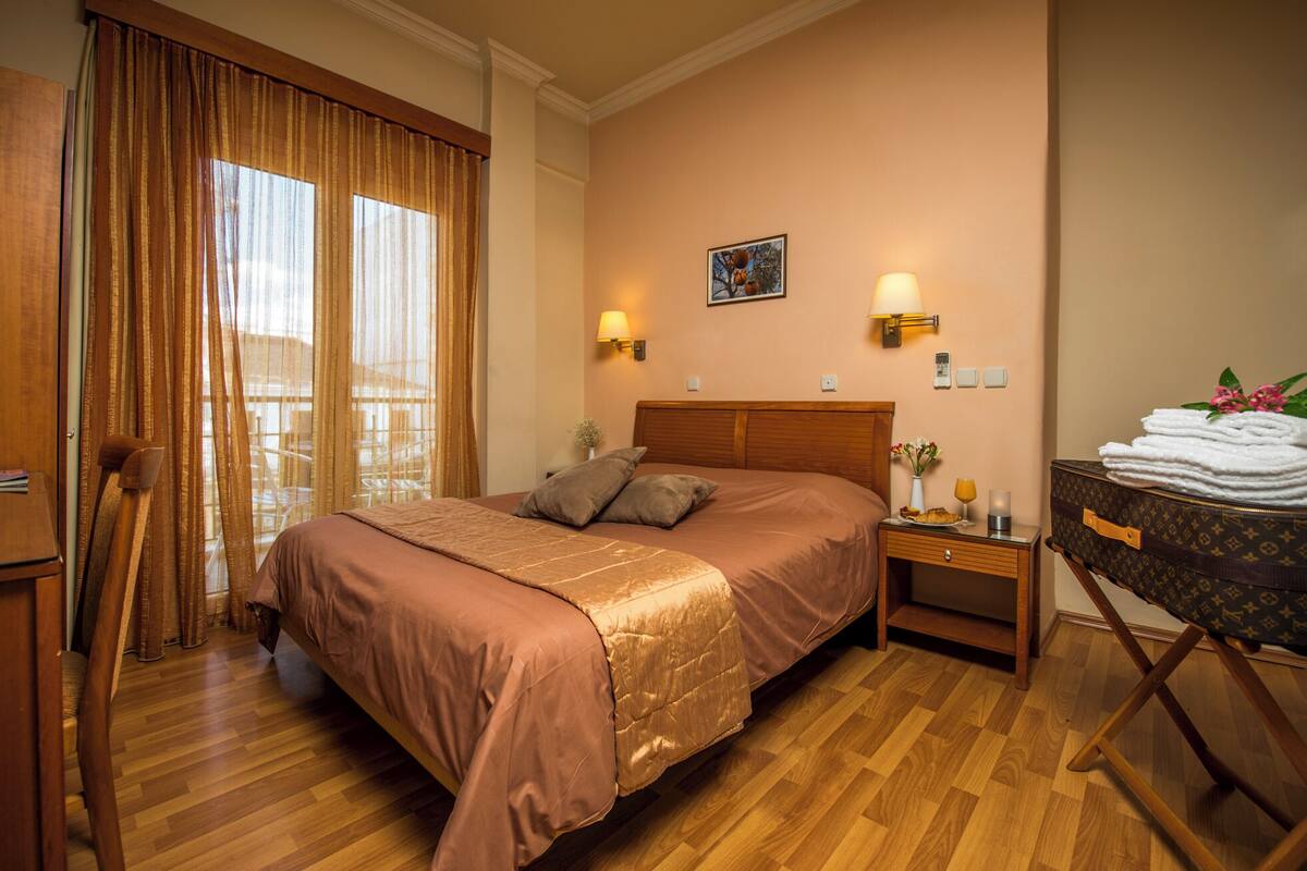 Habitación de hotel con una cama grande, una mesita de noche de madera, una maleta y vistas de la ciudad a través de la ventana.