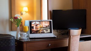 Habitación de hotel con una computadora portátil mostrando el interior del hotel, una mesita de noche con una taza y flores, y un televisor sobre un soporte de madera.