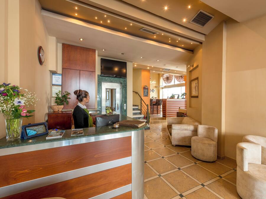 A hotel reception area with a wooden reception desk, a clock, and a vase with flowers. There are armchairs and a staircase in the background.