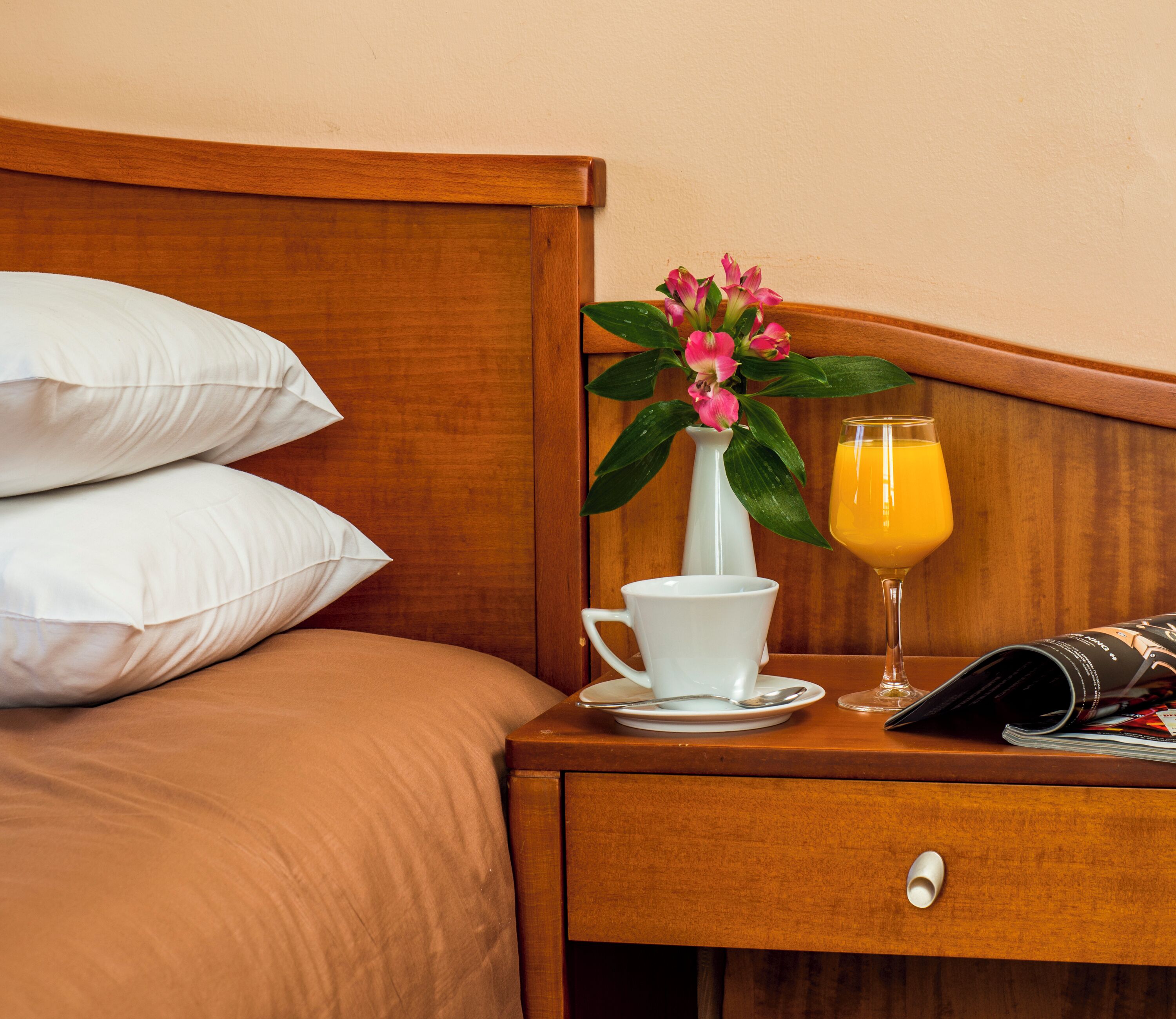 A neatly made bed with white pillows, a wooden headboard, a nightstand with a white cup, a glass of orange juice, and a vase with pink flowers.