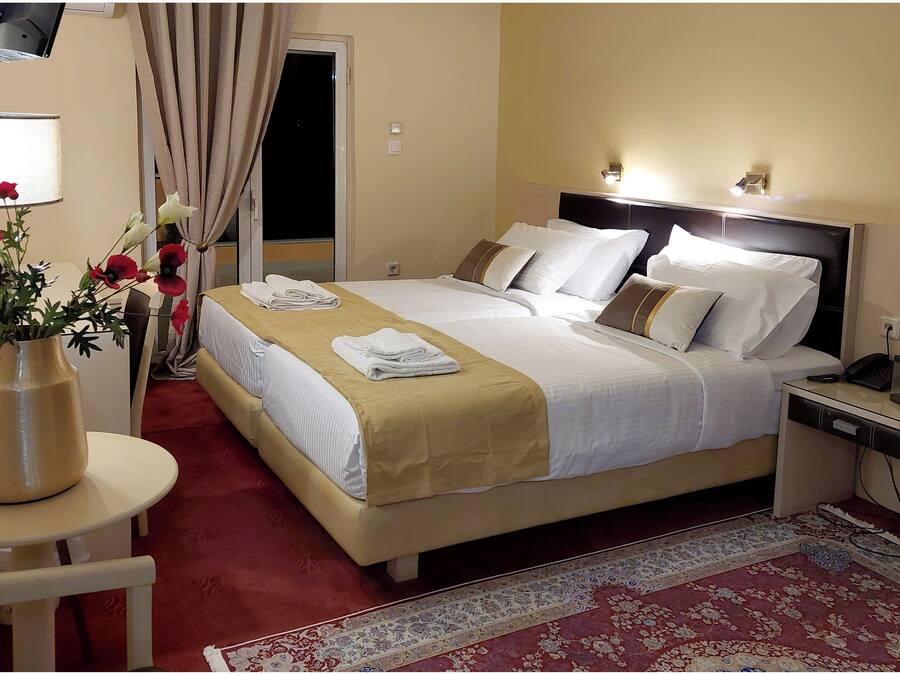 A hotel room with a large bed, a bedside table, a desk, a chair, a vase with flowers, and a window with curtains.