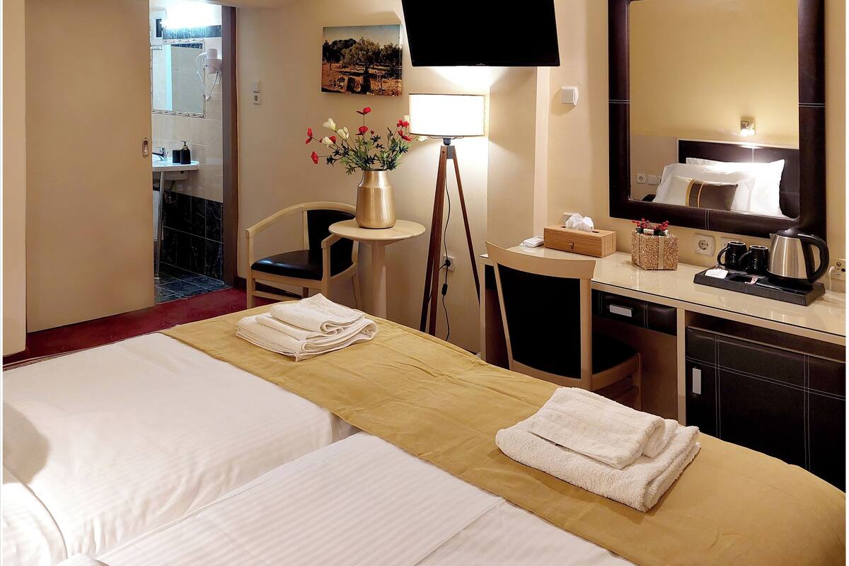 Una habitación de hotel con cama, escritorio, silla, televisión, espejo y un jarrón con flores.