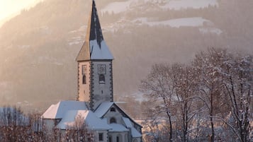 A church with a tall steeple and a clock tower, surrounded by snow-covered trees and a mountainous backdrop.