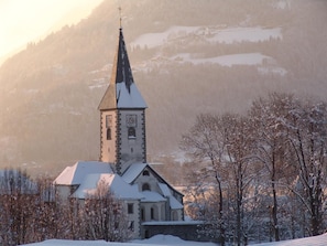 A church with a tall steeple and a clock tower, surrounded by snow-covered trees and a mountainous backdrop. - Gasthof zur Post (Ossiach)