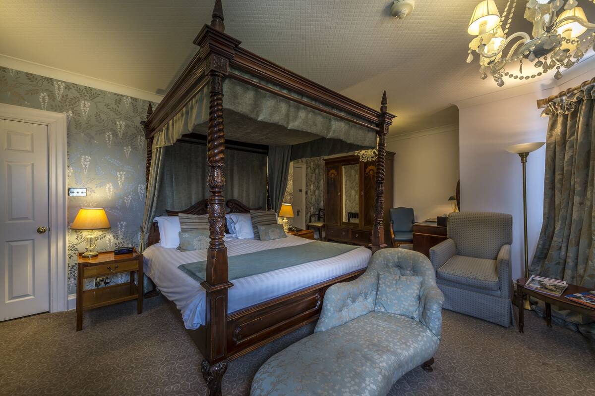 A four-poster bed with a canopy, a sitting chair, a nightstand with a lamp, and a mirror on the wall.