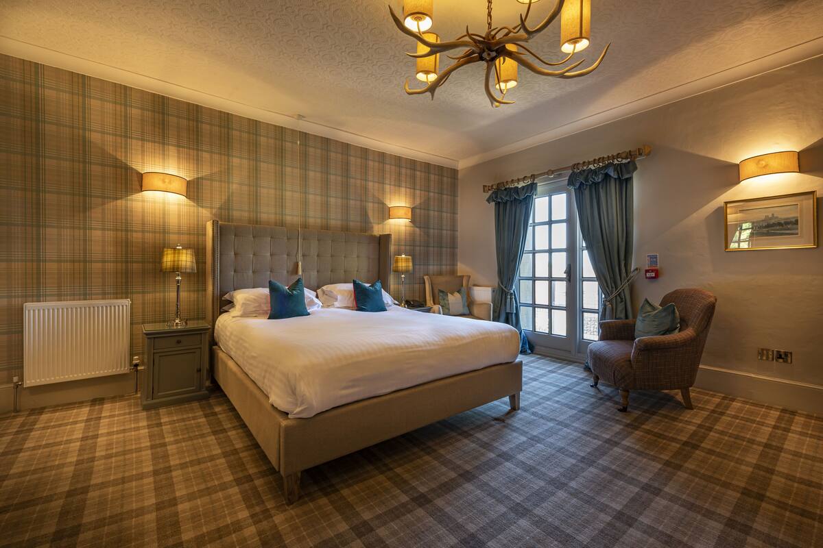 A hotel bedroom with a large bed, two bedside tables, a chair, a window with curtains, and a chandelier.