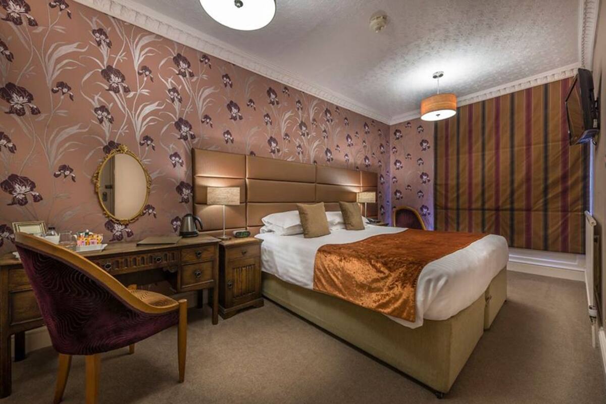A bedroom with a large bed, a desk with a chair, a mirror, and a window with curtains.