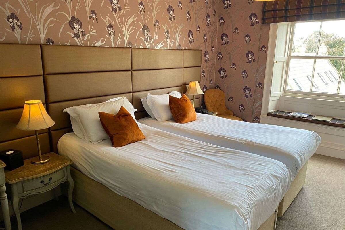 A double bed with white linens and two brown pillows, a bedside table with a lamp, and a window with a view of buildings.