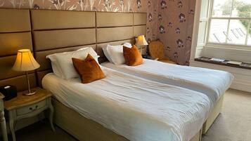 A double bed with white linens and two brown pillows, a bedside table with a lamp, and a window with a view of buildings.