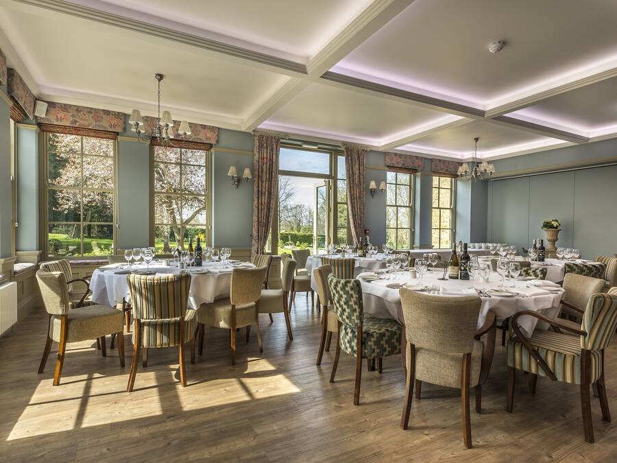 Uma sala de jantar com várias mesas preparadas para refeição, janelas amplas com cortinas e um candelabro.