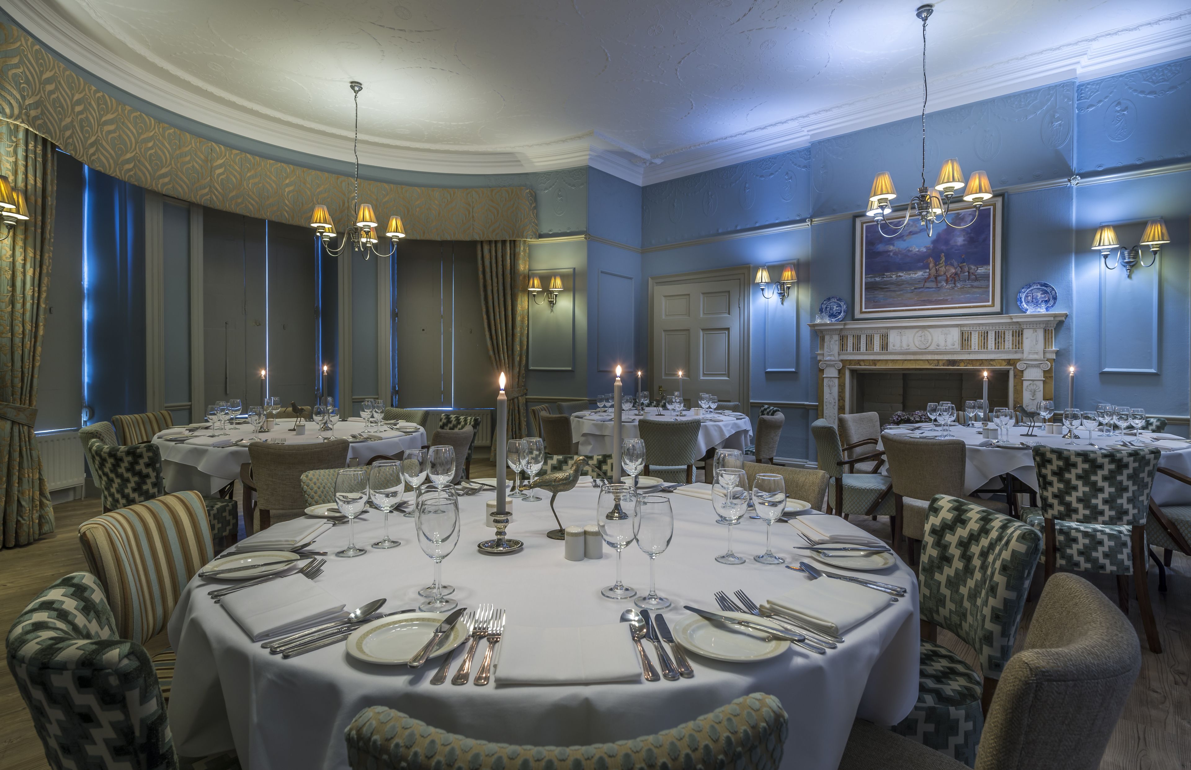 A formal dining room with round tables set for multiple guests, featuring chandeliers and wall sconces.