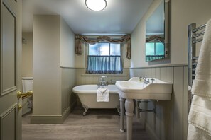Une salle de bain avec une baignoire, deux lavabos, une fenêtre avec des rideaux, un porte-serviettes et une porte dotée d’une poignée en laiton.