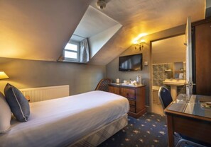 Une chambre à coucher comprenant un lit, un bureau, une chaise, une commode, un téléviseur et une salle de bain visible à travers une porte ouverte.