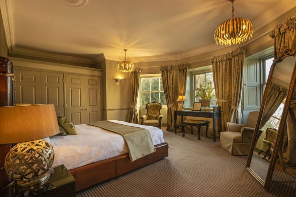 A spacious bedroom with a large bed, a sitting area with a table and chairs, a mirror, and a window with curtains.