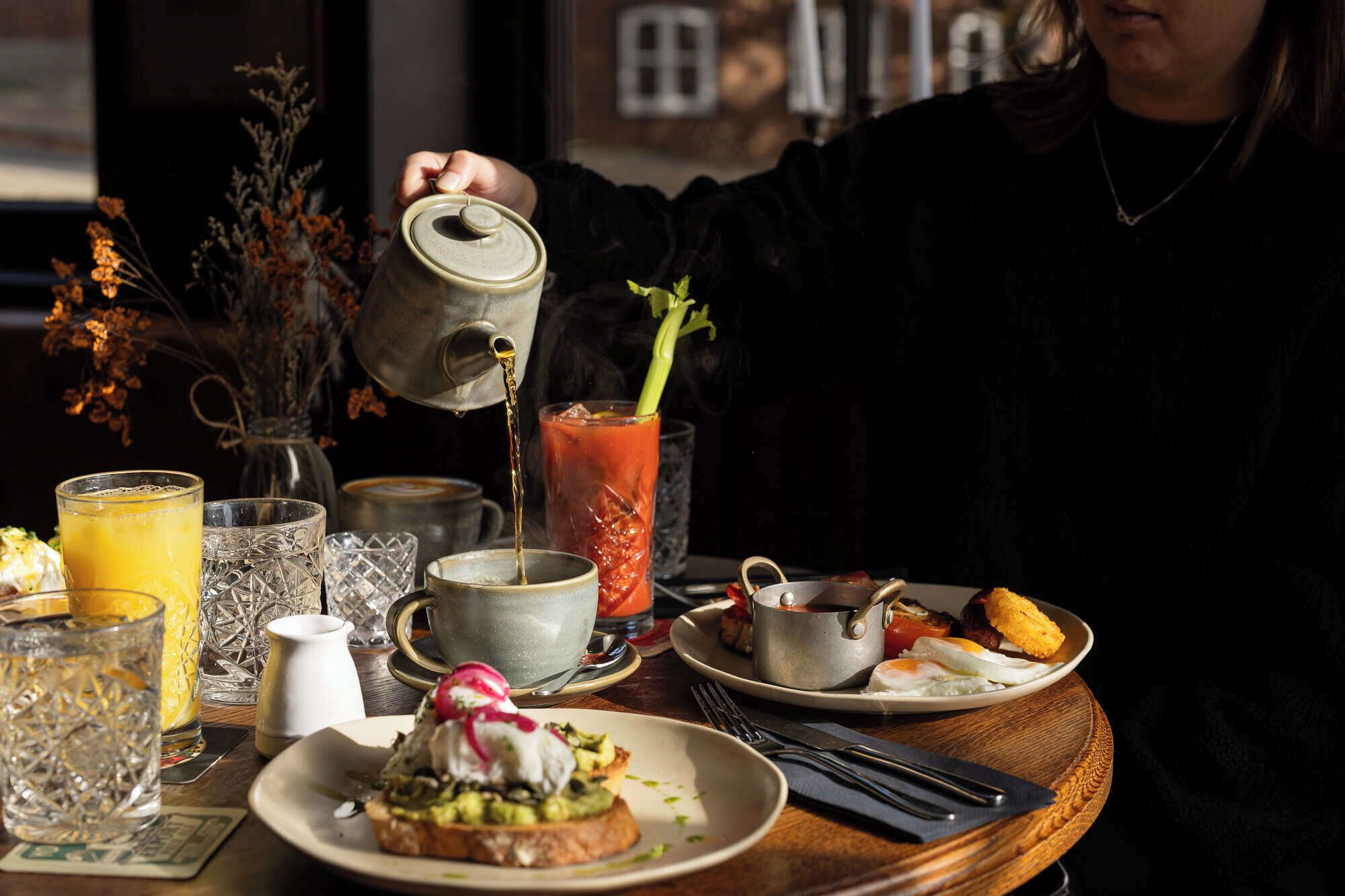 A breakfast table with a variety of dishes, including toast with avocado and a poached egg, a glass of orange juice, and a Bloody Mary.