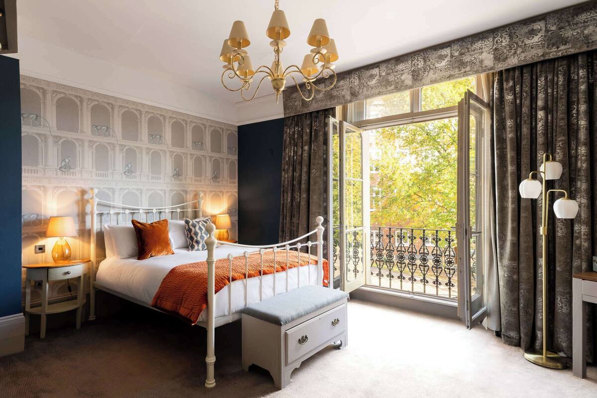 A bedroom with a bed, bedside tables, a bench, a chandelier, and a window with a view.