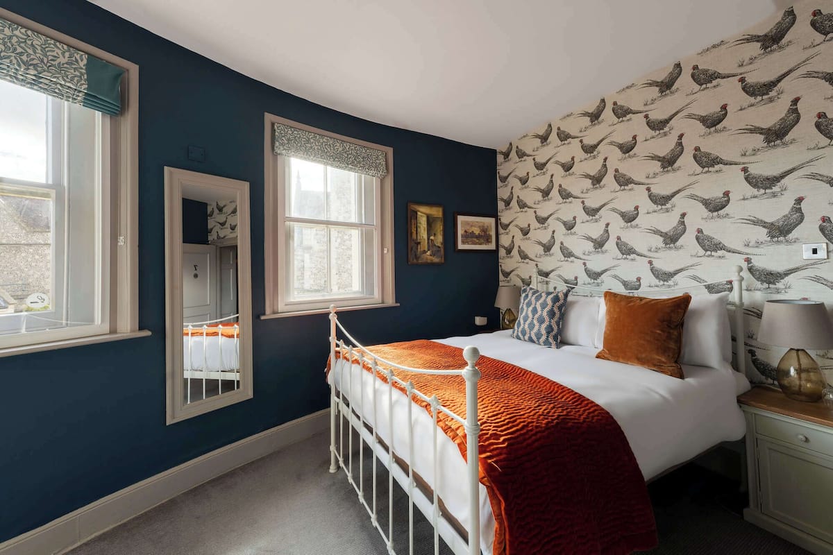 A bedroom with a white bed, orange bedding, a white metal bed frame, a window with a white valance, a mirror, and a wall with a chicken pattern wallpaper.