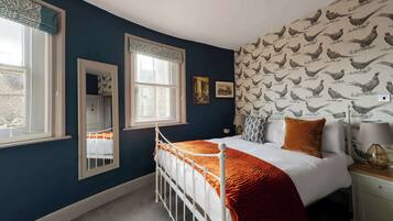Un dormitorio con cama blanca, ropa de cama naranja, estructura de cama metálica blanca, ventana con cortinilla blanca, espejo y pared con papel tapiz estampado con motivos de gallinas.