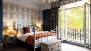 A bedroom with a white four-poster bed, a nightstand with a lamp, a dresser, and a large window with a view of trees. - Westgate Winchester (Winchester)
