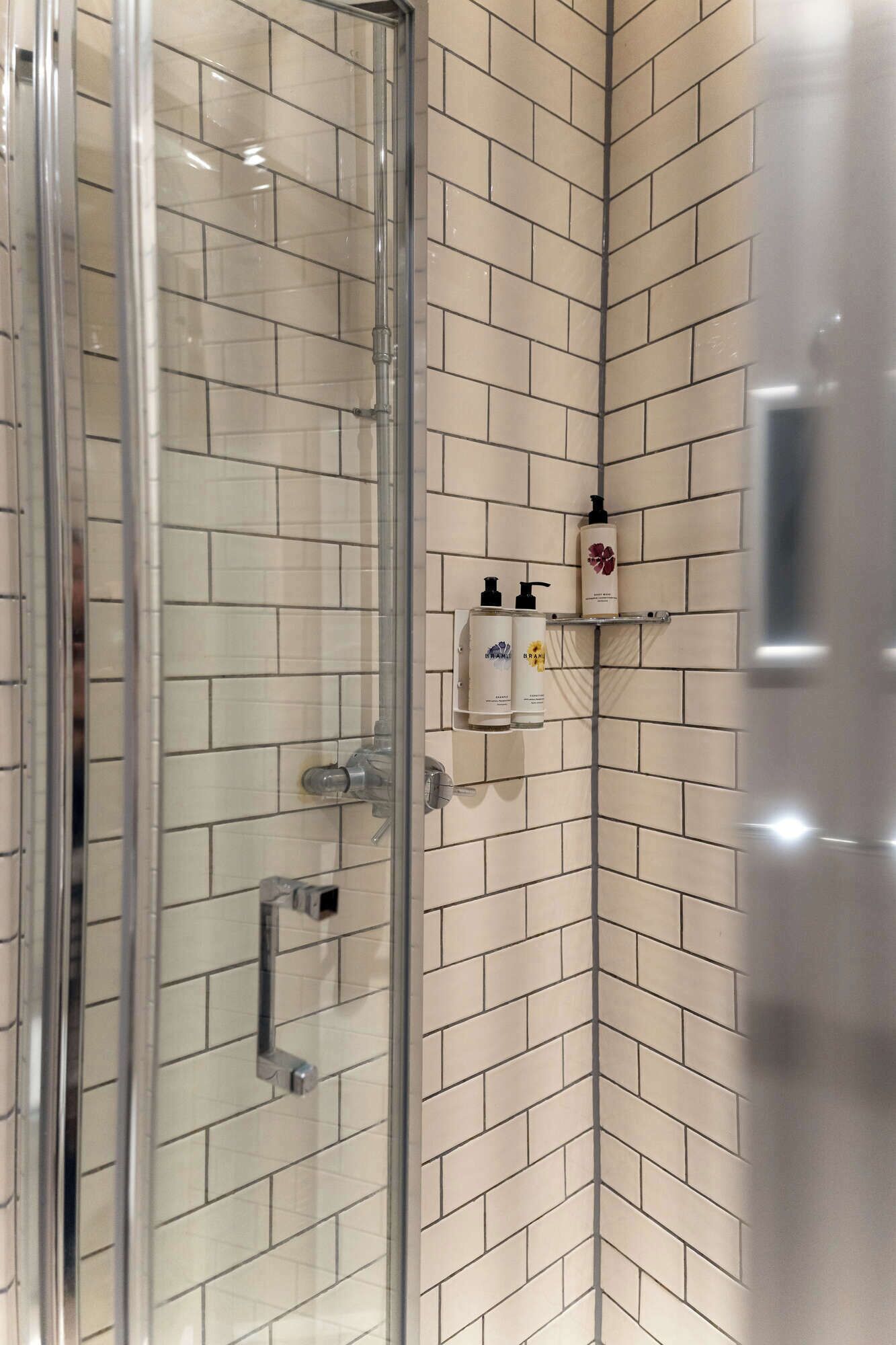 Douchegedeelte met een glazen deur, betegelde wanden en twee shampoo flessen op een plank.