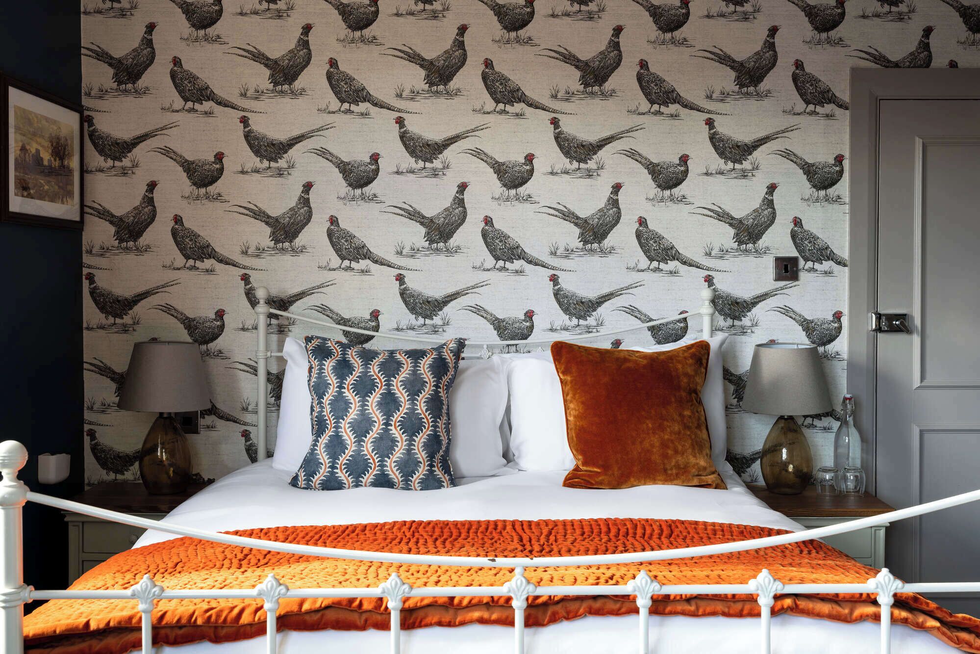 A bedroom with a patterned wallpaper, a white bed with an orange blanket, and two bedside lamps.