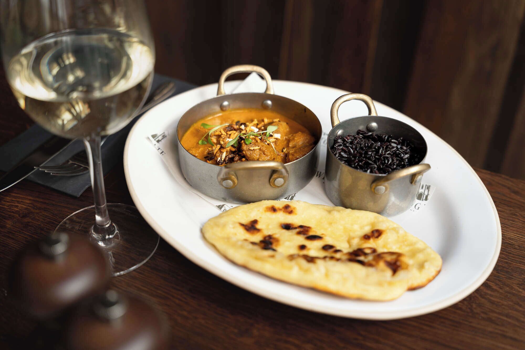 Ein Teller mit Curry, schwarzem Reis und Naan-Brot, dazu ein Glas Weißwein.