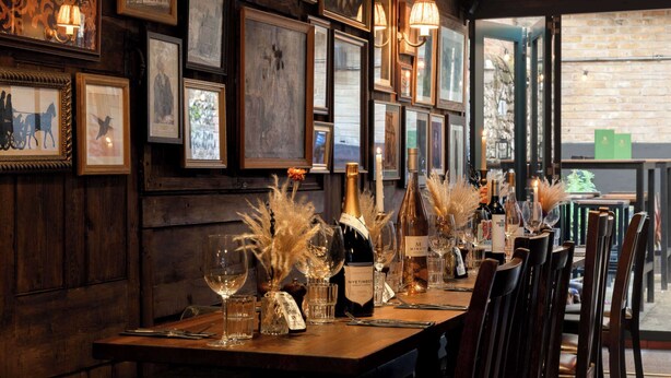 A restaurant with a long wooden table set for dining, framed pictures on the wall, and a view of the outside through large windows. - Westgate Winchester (Winchester)